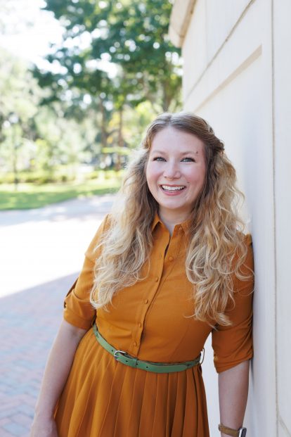 Jessica-Jean smiling in an orange dress with a green belt leaning against a white wall, with greenery in the background