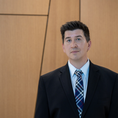 “Professor at the University of Florida wearing a dark pinstripe suit, white dress shirt, and plaid tie, standing in front of a modern wooden wall with geometric paneling.