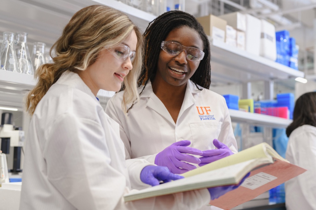 Two people wearing lab coats and safety glasses review a notebook in a laboratory setting