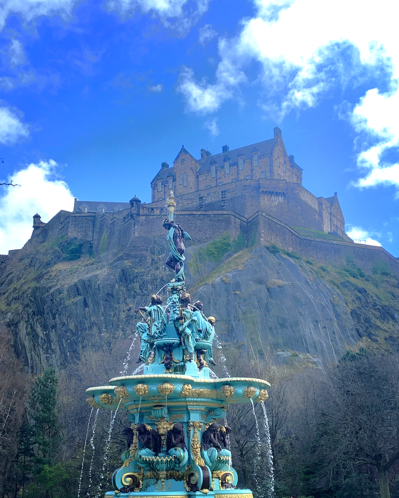 Ornate multi-tiered fountain in the foreground with a large stone castle perched atop a steep rocky cliff in the background